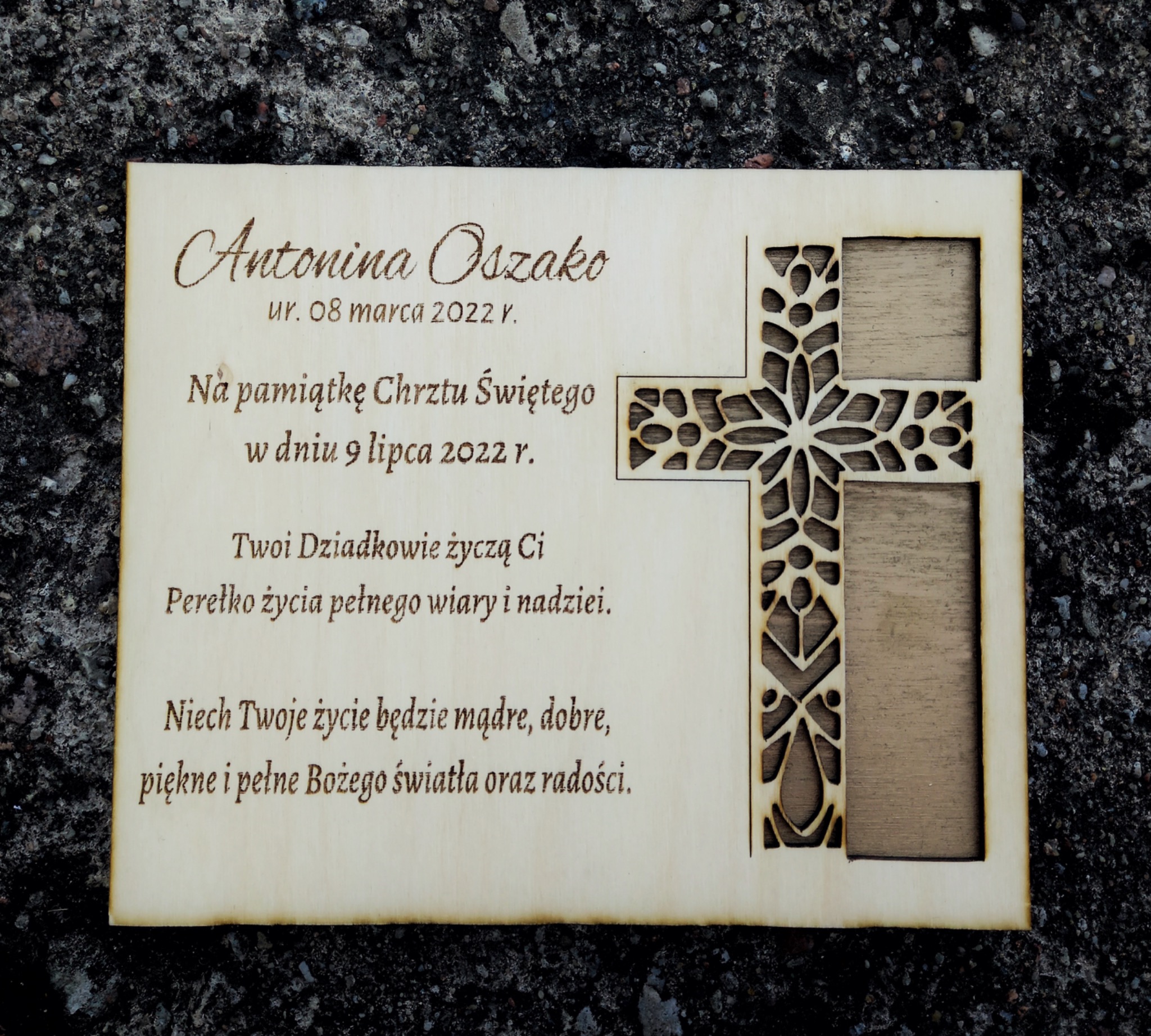 Drewniana pamiątka Chrztu Świętego z wygrawerowanym imieniem Antonina Oszako, datą urodzenia i życzeniami od dziadków, ozdobiona ażurowym krzyżem, leżąca na szarym, kamiennym tle.