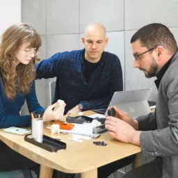 Zespół trzech osób przy biurku analizuje modele 3D wydrukowane z drukarki 3D, widoczny laptop i narzędzia pomiarowe.