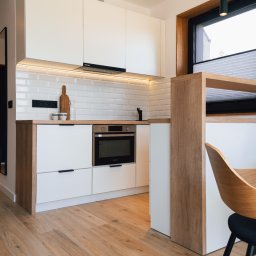 Grid Studio Projektowe - Nowoczesna kuchnia z białymi szafkami i drewnianymi akcentami, oświetleniem LED pod szafkami wiszącymi, drewnianą deską do krojenia opartą o ścianę z białych płytek, oraz fragmentem krzesła...