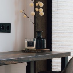 Grid Studio Projektowe - Dekoracyjny kącik: Ciemny drewniany blat biurka z czarnym gniazdkiem elektrycznym, świecą zapachową 'Home Fragrance' na drewnianym plastrze, ciemnym wazonem z gałązką z kremowymi kulkami, na tle...