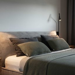 Grid Studio Projektowe - Minimalistyczna sypialnia z szarym tapicerowanym łóżkiem, drewnianą szafką nocną i czarnymi lampkami ściennymi