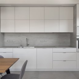 Grid Studio Projektowe - Minimalistyczna kuchnia z białymi szafkami, szarym blatem i backsplashiem imitującym beton. Na pierwszym planie fragment drewnianego stołu i szare krzesła.