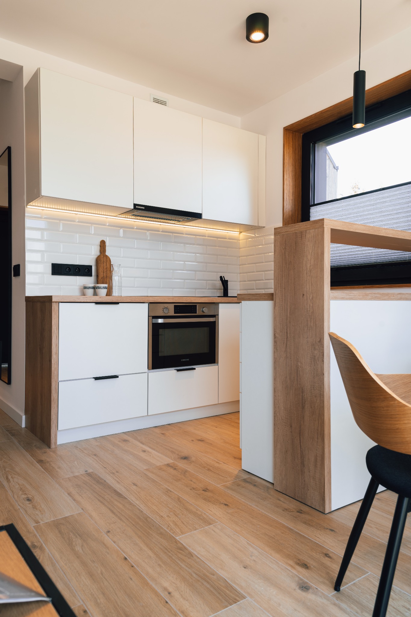 Nowoczesna kuchnia z białymi szafkami i drewnianymi akcentami, oświetleniem LED pod szafkami wiszącymi, drewnianą deską do krojenia opartą o ścianę z białych płytek, oraz fragmentem krzesła...