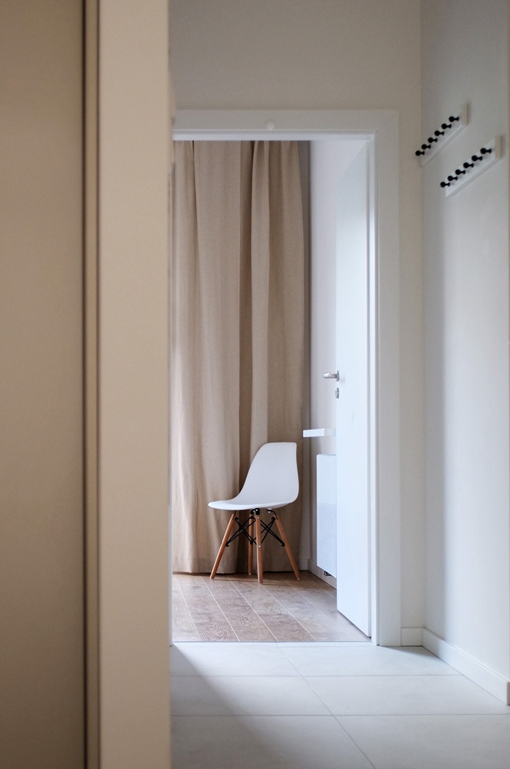 Minimalistyczne wnętrze z białym krzesłem Eames na drewnianej podłodze, widoczne przez otwarte drzwi z białymi ścianami i beżową zasłoną w tle, oświetlone naturalnym światłem.