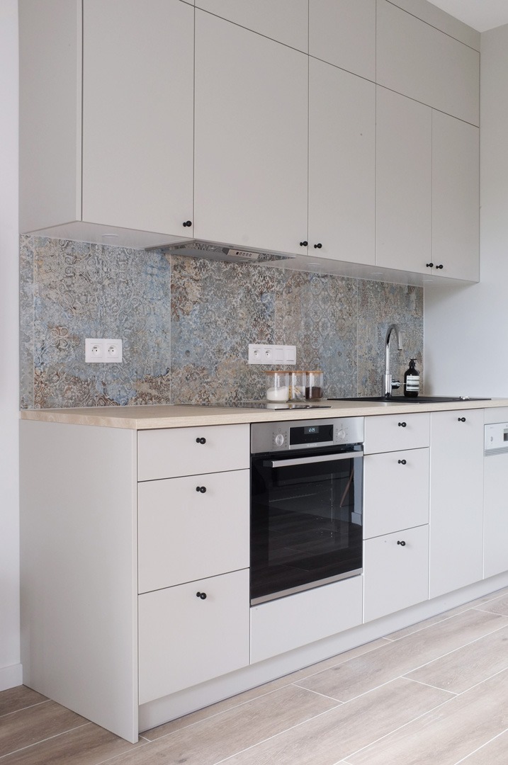 Jasna kuchnia z białymi szafkami, czarnymi gałkami, wzorzystą płytą nad blatem i drewnopodobną podłogą. Na blacie słoiki z produktami spożywczymi.