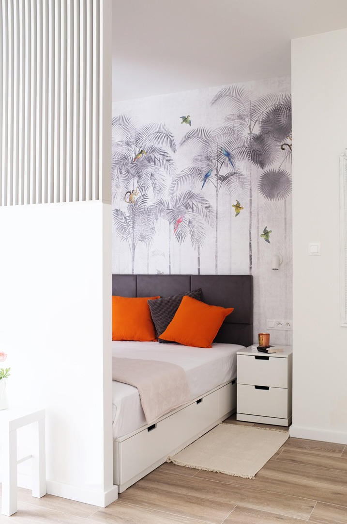 Sypialnia z łóżkiem z szufladami, pomarańczowymi poduszkami i tapetą w tropikalne drzewa, widok zza białej ścianki działowej z pionowymi listwami.