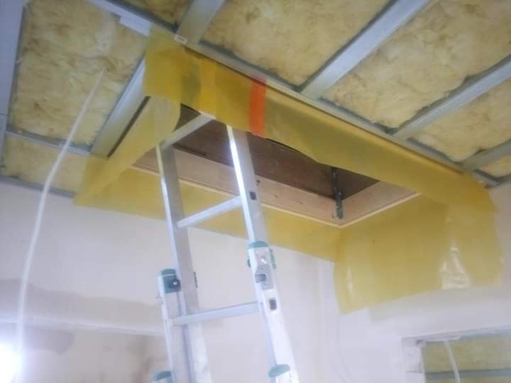 Poddasze w trakcie izolacji, widoczna wełna mineralna między metalowymi profilami, zabezpieczona żółtą folią malarską wokół otworu włazowego, drabina aluminiowa oparta o ścianę.