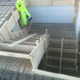 Konstrukcja szalunku schodów zbrojonych w trakcie budowy, widoczna siatka zbrojeniowa, ściany z bloczków oraz odblaskowa kurtka robocza.