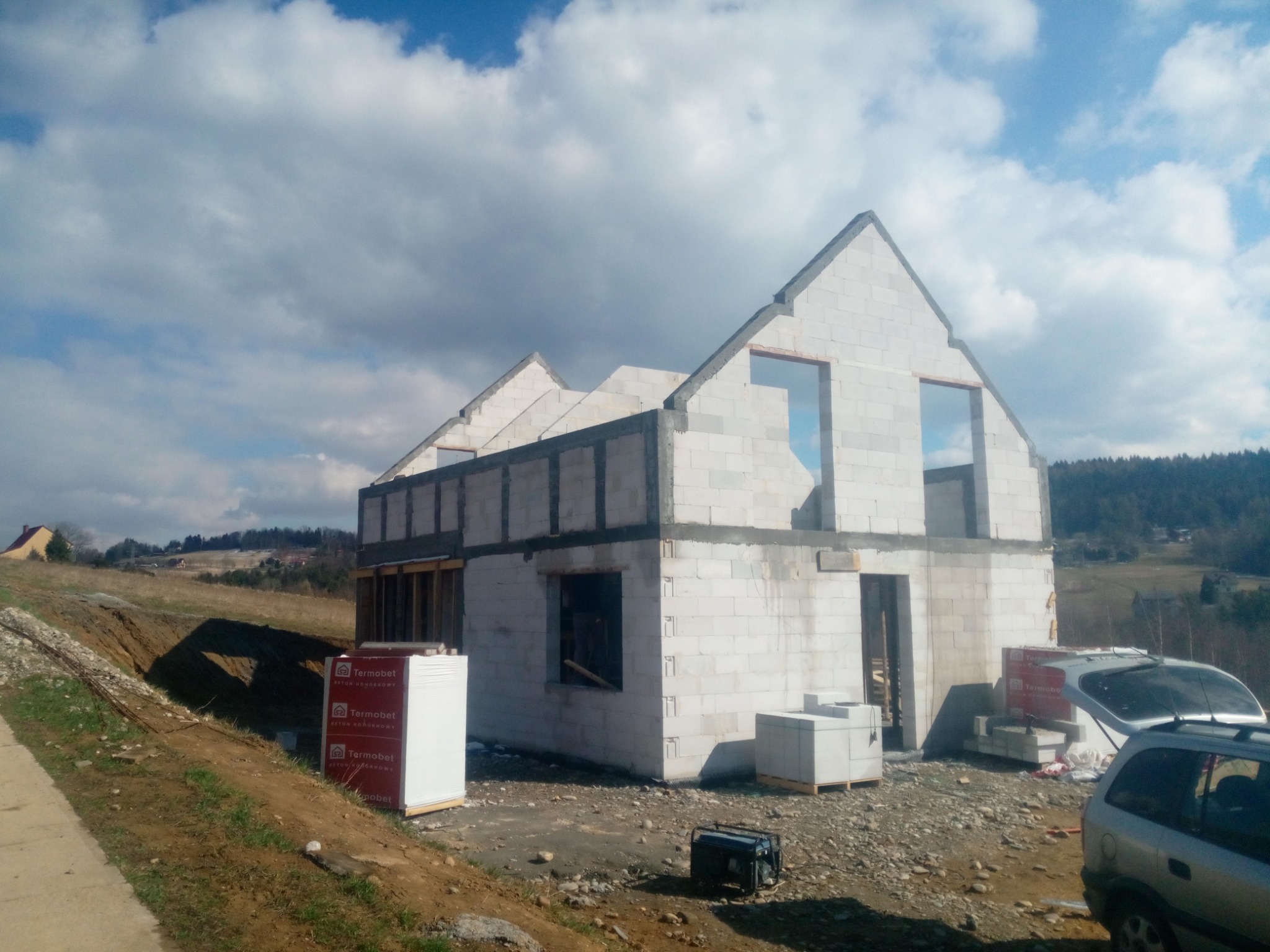 Surowy stan domu w trakcie budowy, widoczne ściany z pustaków, otwory okienne bez ram, paleta z materiałami budowlanymi Termobet i generator prądu na placu budowy.