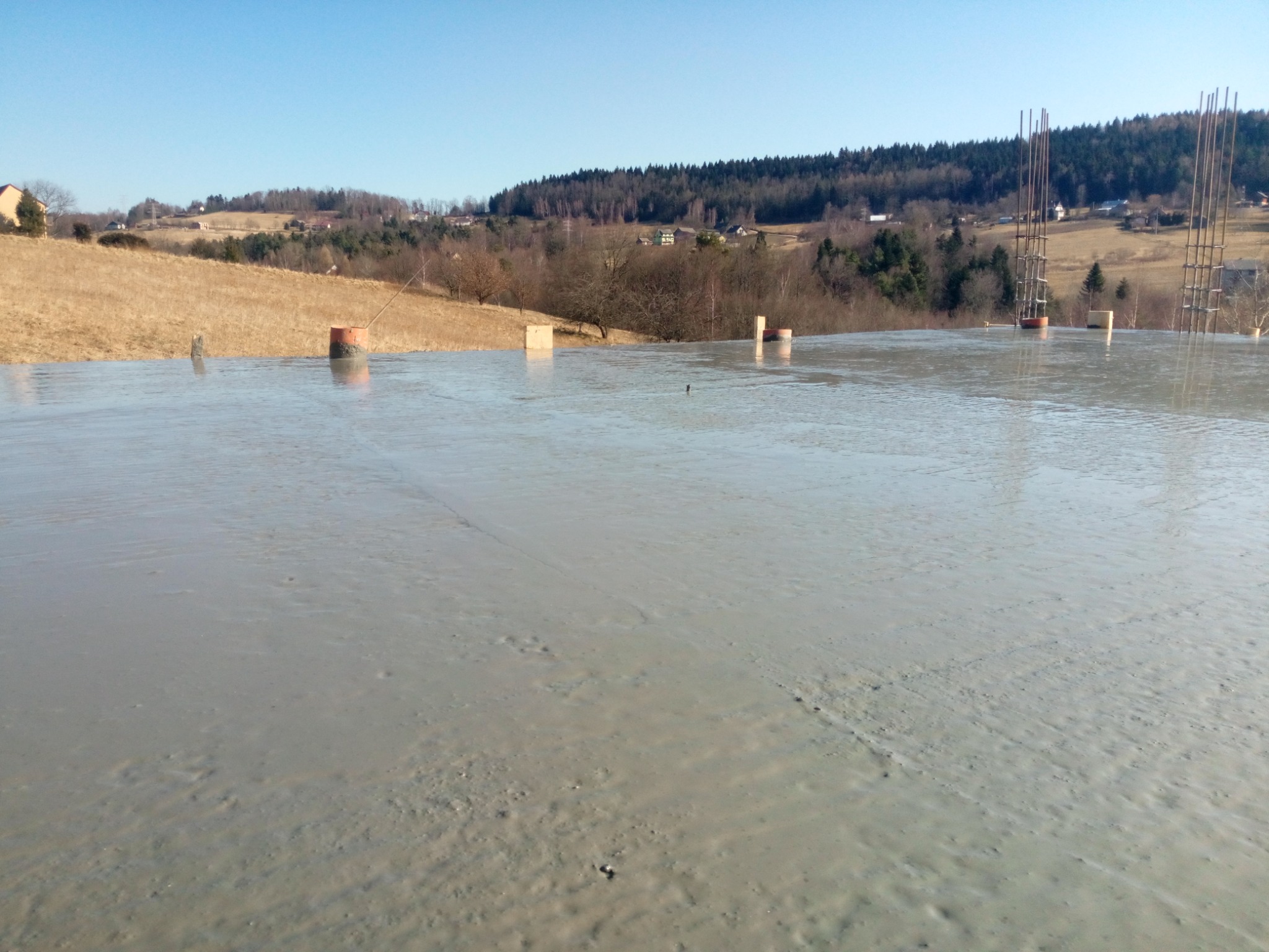Świeżo wylany, mokry strop betonowy z wystającymi prętami zbrojeniowymi na tle pagórkowatego krajobrazu z lasem i zabudowaniami w oddali.