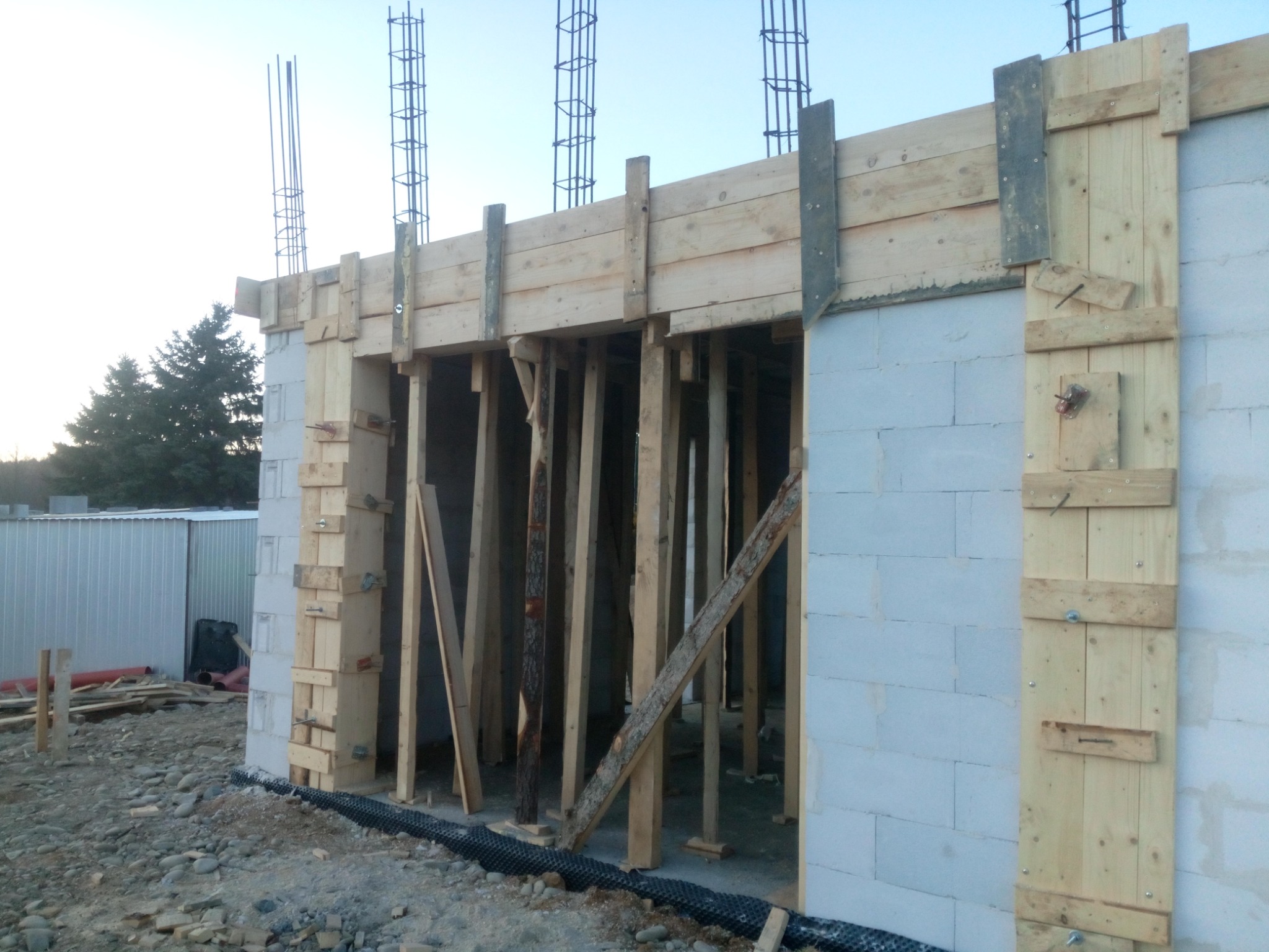 Budowa domu z bloczków betonowych, widoczne drewniane szalunki i zbrojenia pionowe wystające ponad konstrukcję, w tle drzewa i metalowe ogrodzenie.