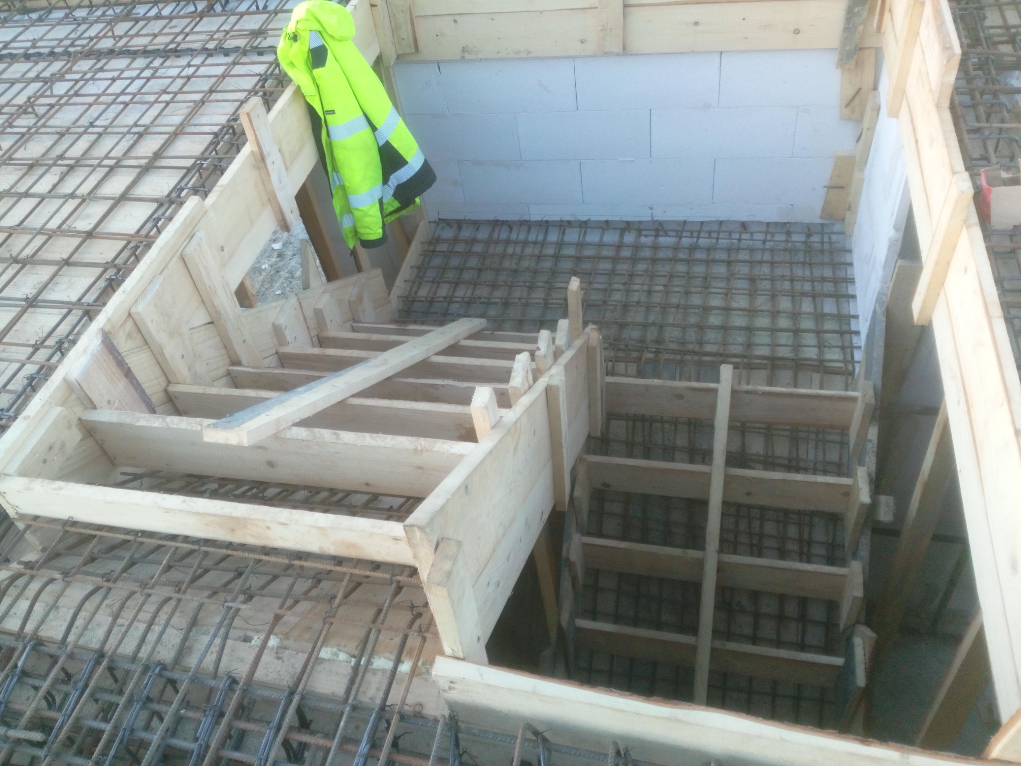 Konstrukcja szalunku schodów zbrojonych w trakcie budowy, widoczna siatka zbrojeniowa, ściany z bloczków oraz odblaskowa kurtka robocza.