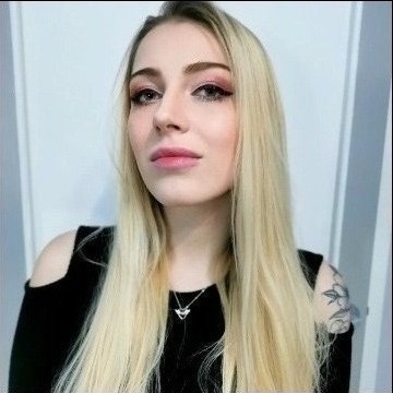 Portret kobiety z blond włosami i delikatnym, różowym odcieniem na powiekach oraz ustach, ubrana w czarną bluzkę z odkrytymi ramionami i srebrny naszyjnik, na ramieniu widoczny tatuaż.