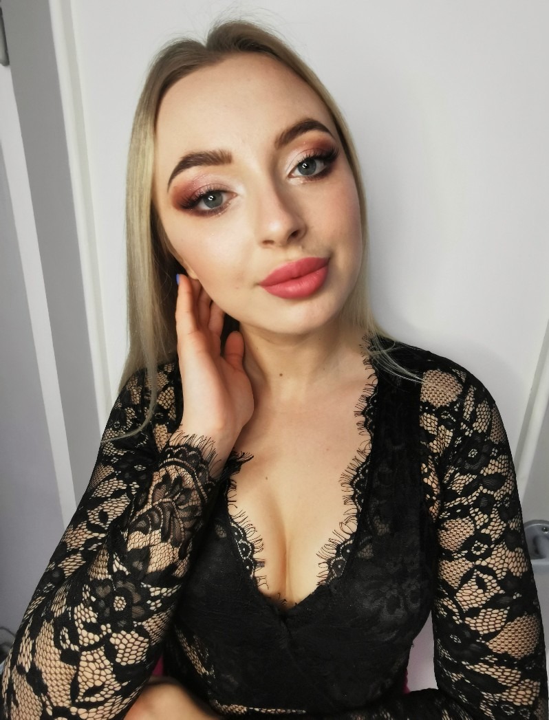 Portret blondynki w czarnej koronkowej sukience, z brązowo-różowym cieniowaniem powiek i matową pomadką w kolorze dusty rose, delikatnie dotykającej policzka palcami z niebieskim lakierem.