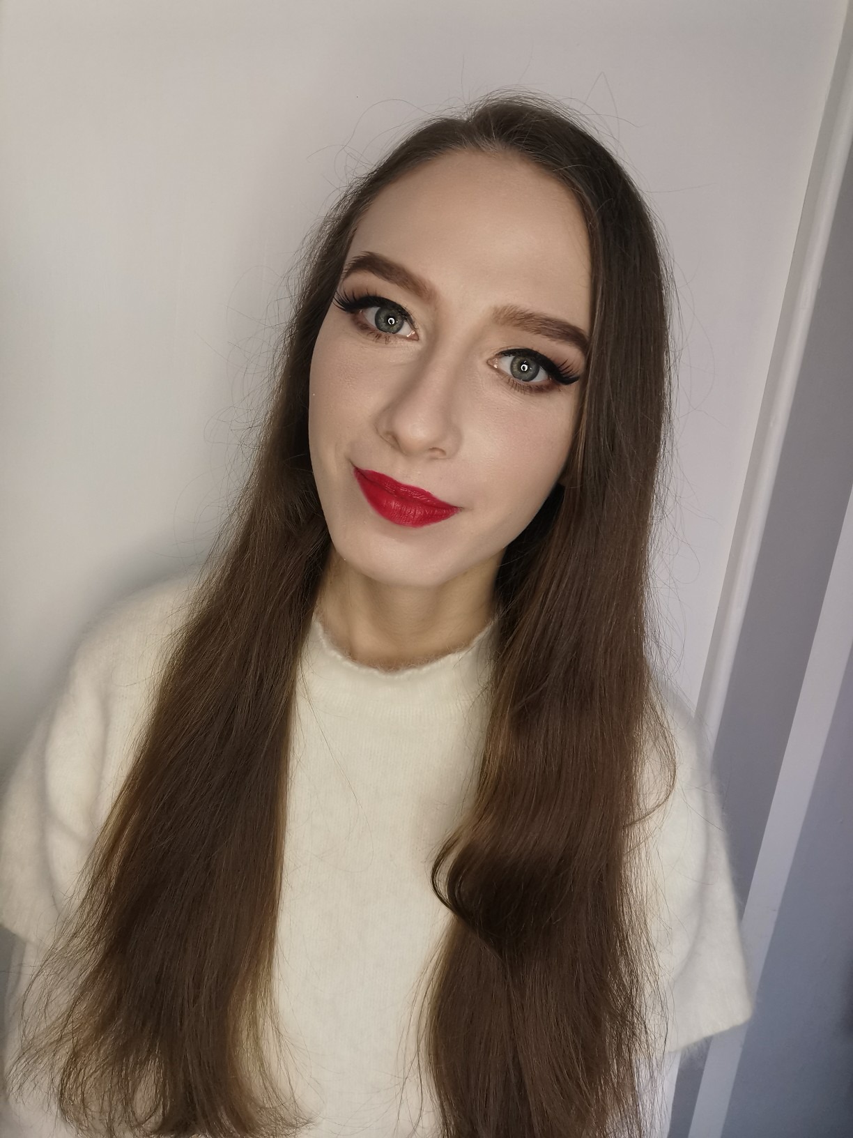 Portret kobiety z długimi brązowymi włosami, podkreślonymi oczami z czarnym eyelinerem i intensywnie czerwoną szminką na ustach, ubrana w kremowy sweter.