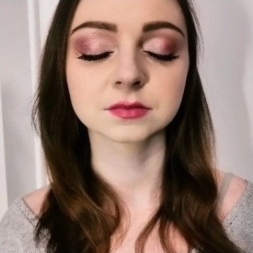 Portret kobiety z zamkniętymi oczami, prezentującej subtelny, różowy cień do powiek z delikatnym połyskiem i precyzyjną kreską eyelinerem oraz pomalowane usta błyszczącą pomadką w odcieniu fuksji.