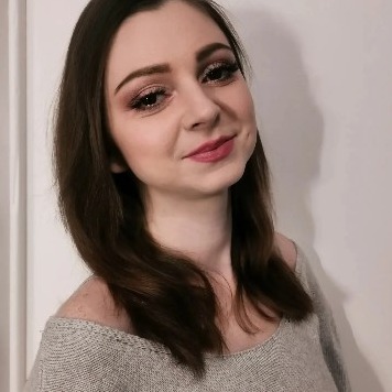 Portret kobiety z subtelnym, wieczorowym upiększeniem oczu i ust, delikatny rumieniec, ciemne włosy opadające na ramiona, uśmiech z dołeczkiem w policzku, szary sweter na tle białej ściany.