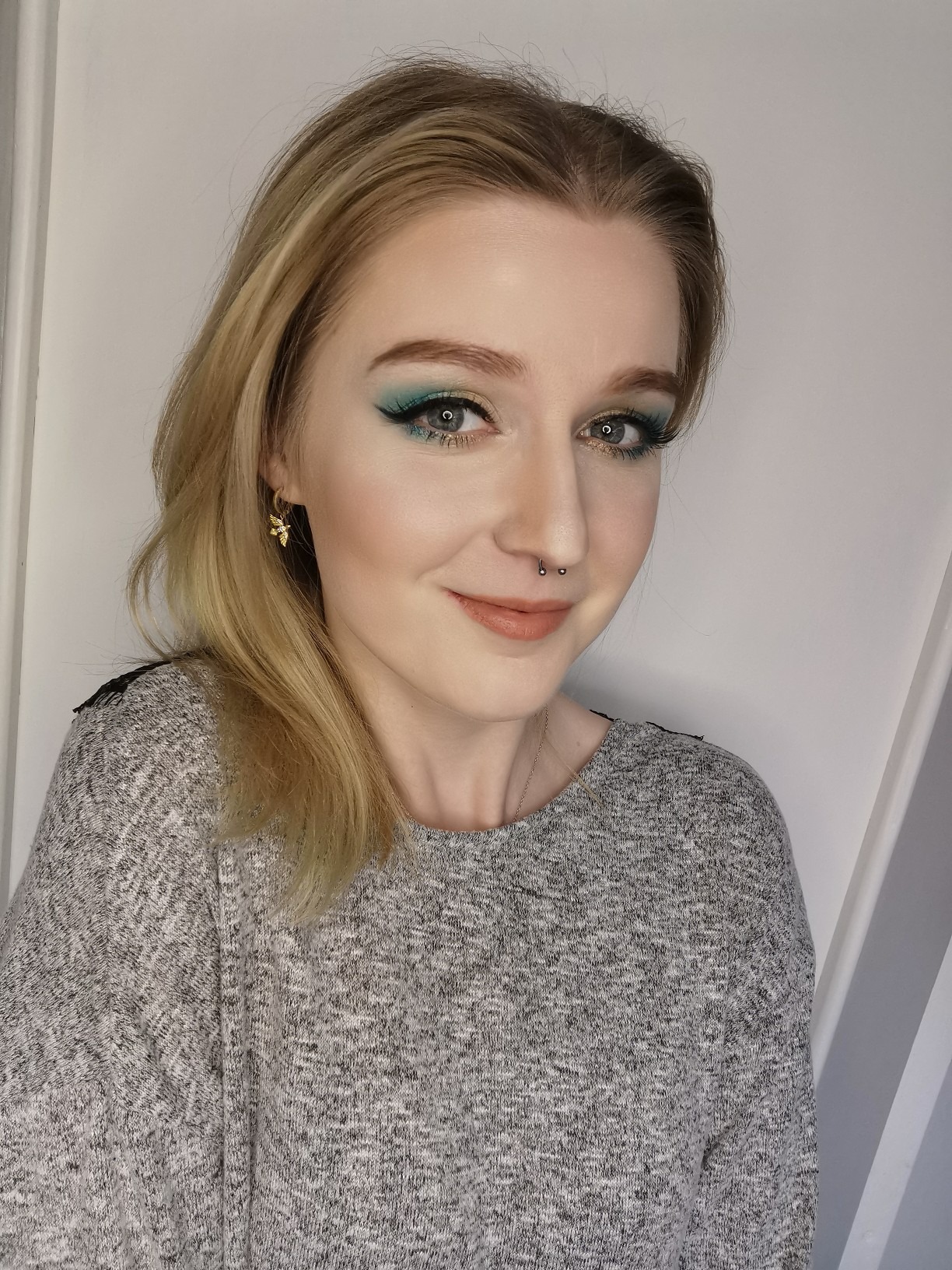 Portret uśmiechniętej blondynki z intensywnym turkusowym cieniem na powiekach, delikatnym konturowaniem twarzy i kolczykami w kształcie pszczółek.