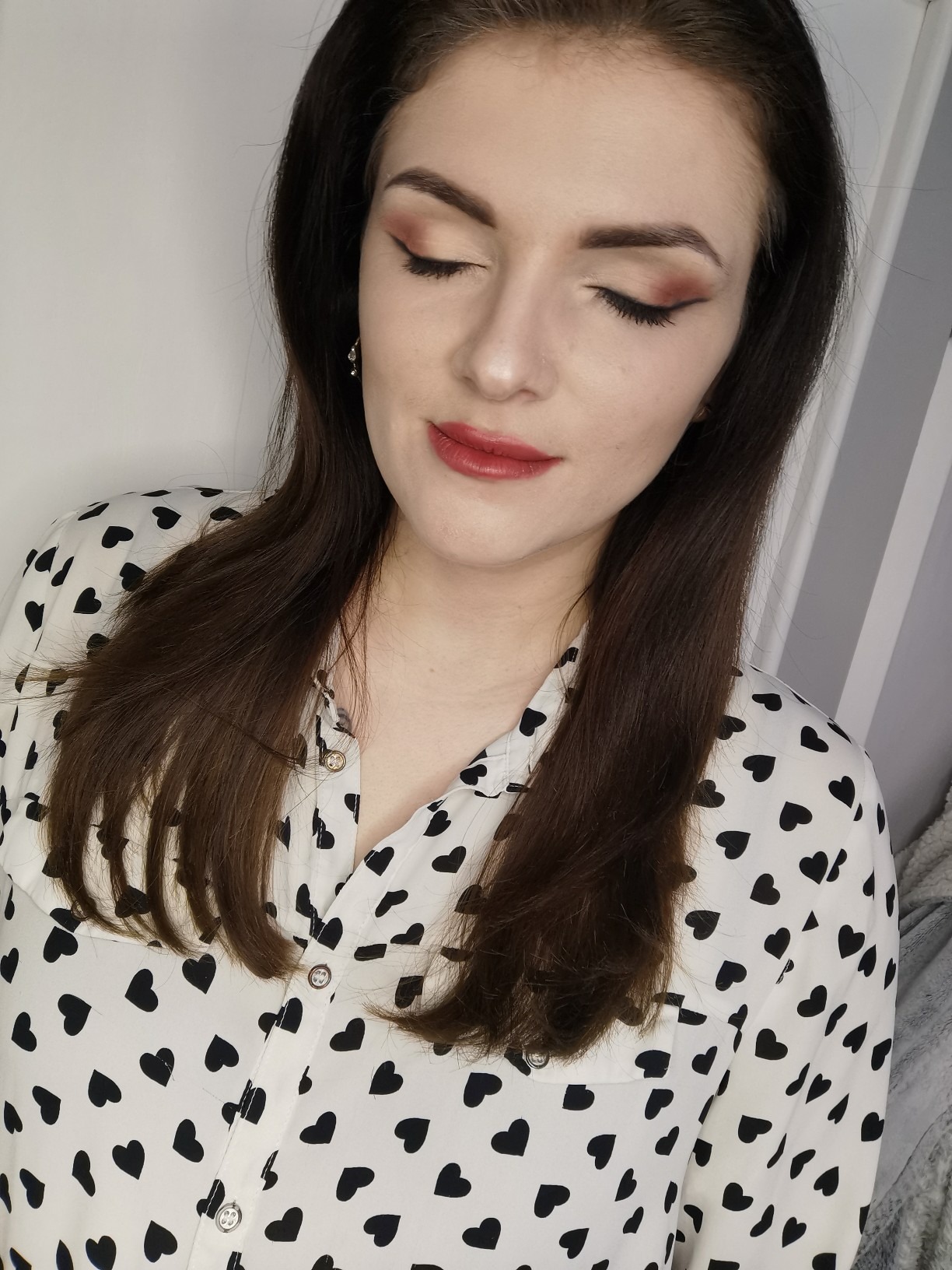 Portret kobiety z zamkniętymi oczami prezentującej delikatny, dzienny make-up z brązowymi cieniami i czerwoną pomadką, ubrana w koszulę w czarne serca.