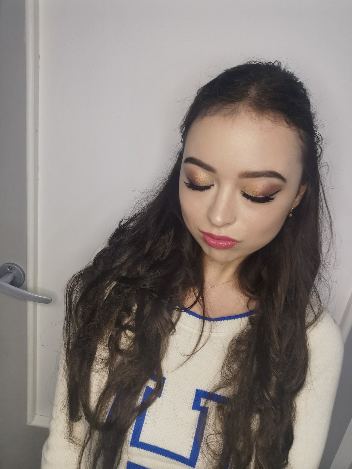 Portret kobiety z brązowym makijażem oczu i różowymi ustami, patrzącej w dół, z widocznymi długimi, kręconymi włosami i fragmentem białego swetra z niebieskim obramowaniem.