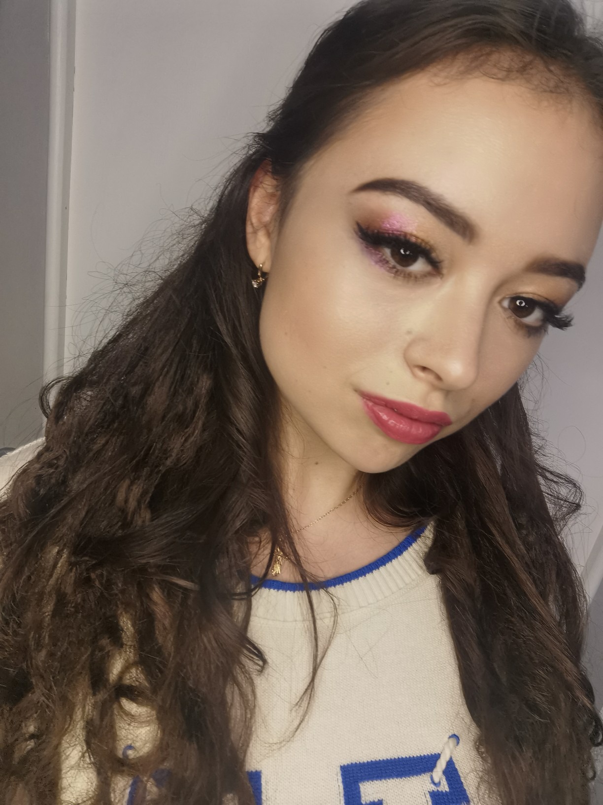 Portret kobiety z błyszczącym, różowym cieniem na powiekach i mocno podkreślonymi ustami, delikatny konturing twarzy, włosy częściowo upięte, złota biżuteria.