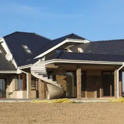 Dom jednorodzinny w trakcie budowy, z ciemną dachówką, widoczne rynny, okna dachowe, komin, schody spiralne i elementy wykończeniowe elewacji.