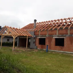 Surowa konstrukcja domu w trakcie budowy: widoczna murowana ściana z otworami okiennymi i drewniana więźba dachowa oraz zadaszenie tarasu w stanie surowym, na tle pochmurnego nieba.