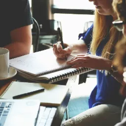 Grupa osób przy drewnianym stole, notatnik z rysunkami, laptop, filiżanka kawy, analiza dokumentów w jasnym pomieszczeniu.