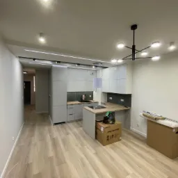 Nowoczesna, jasna kuchnia z szarymi szafkami, drewnianym blatem i parkietem; widoczne kartony po sprzęcie AGD i nowoczesne oświetlenie.