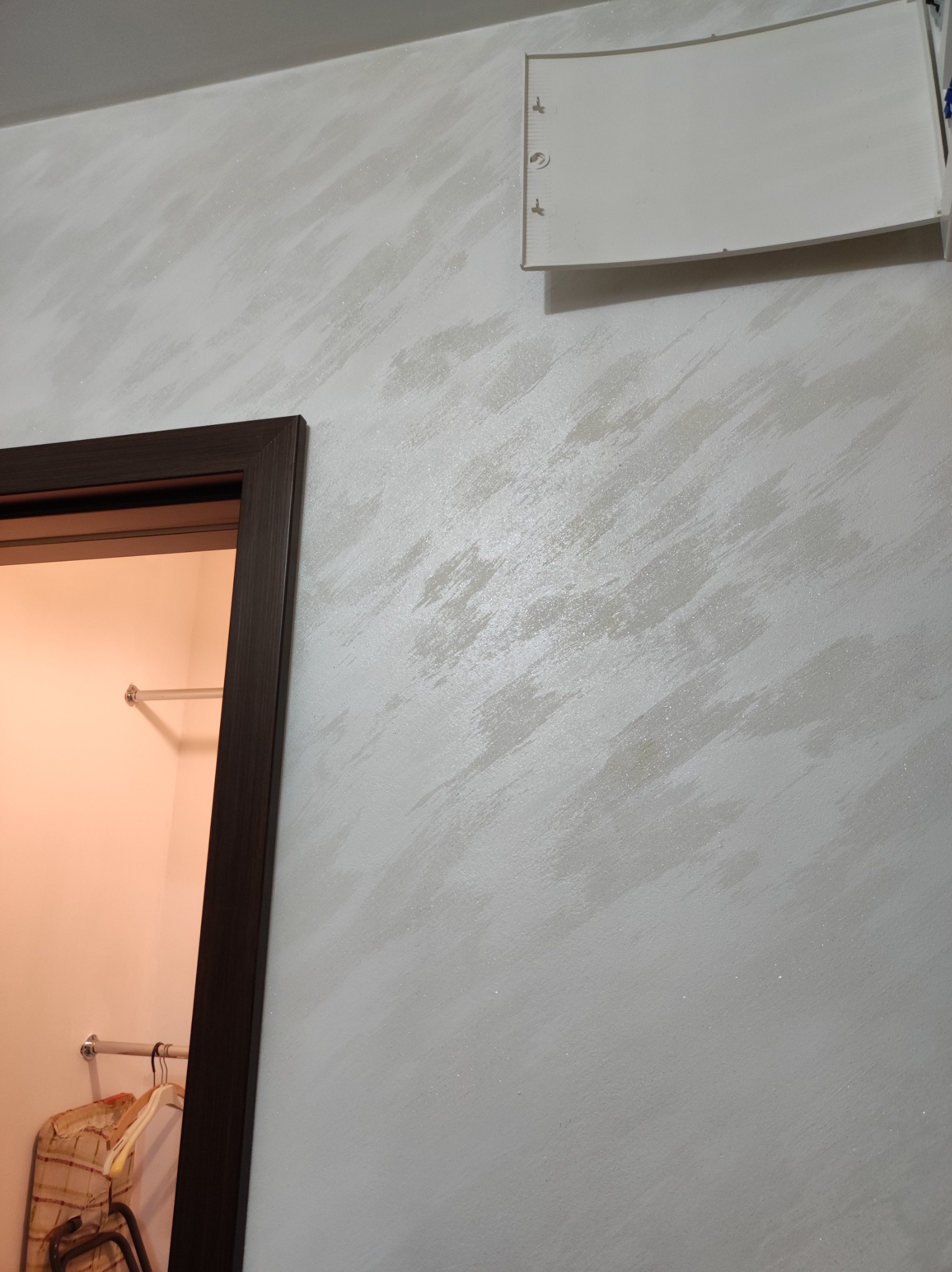 Ściana z dekoracyjną, jasną powłoką malarską z efektem smug, widoczny fragment futryny drzwi i biały element instalacji na ścianie.
