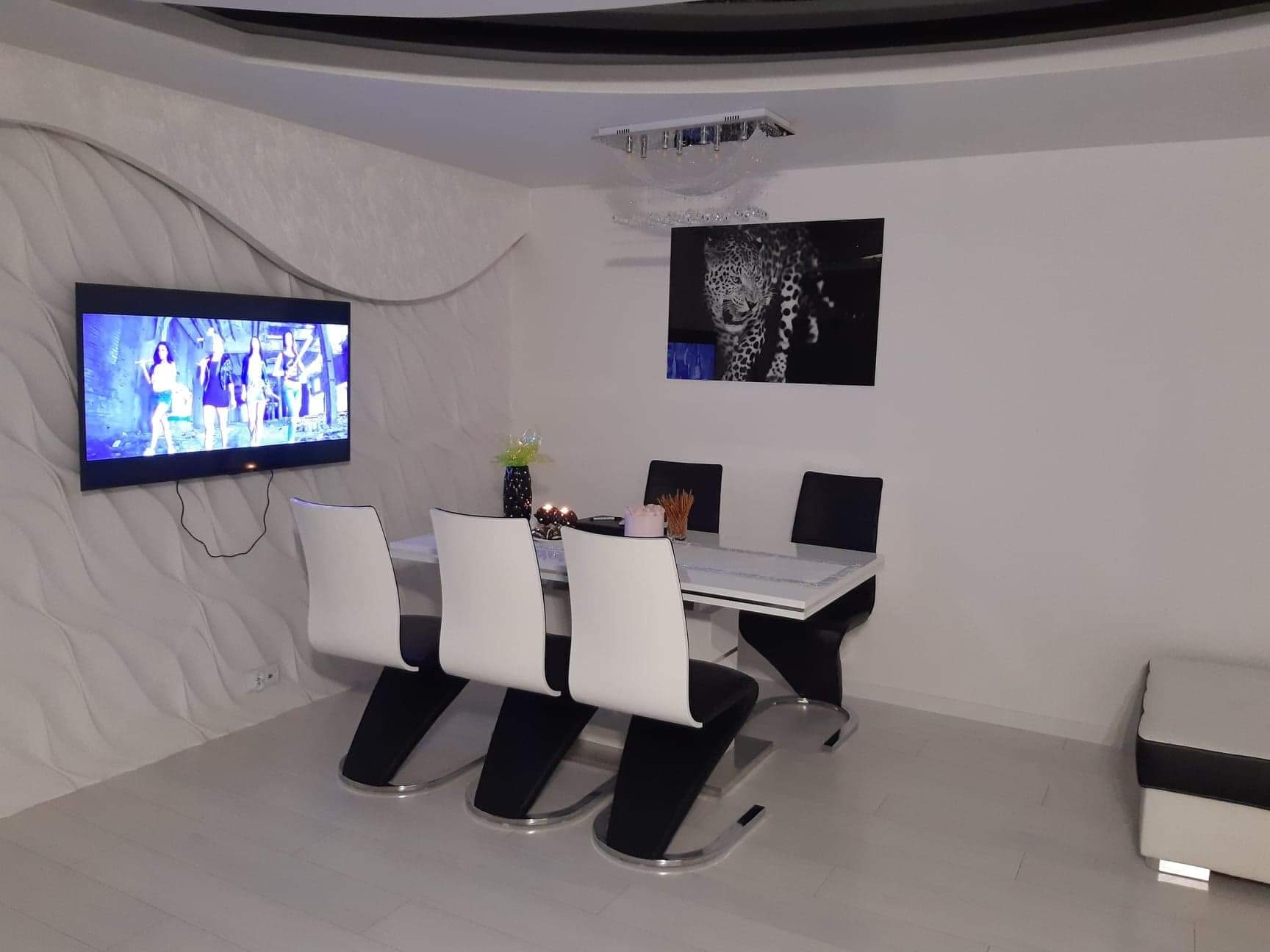 Nowoczesne wnętrze jadalni z białą, teksturowaną ścianą i telewizorem, biały stół z czterema krzesłami, czarno-biały obraz z motywem zwierzęcym, designerska lampa sufitowa.