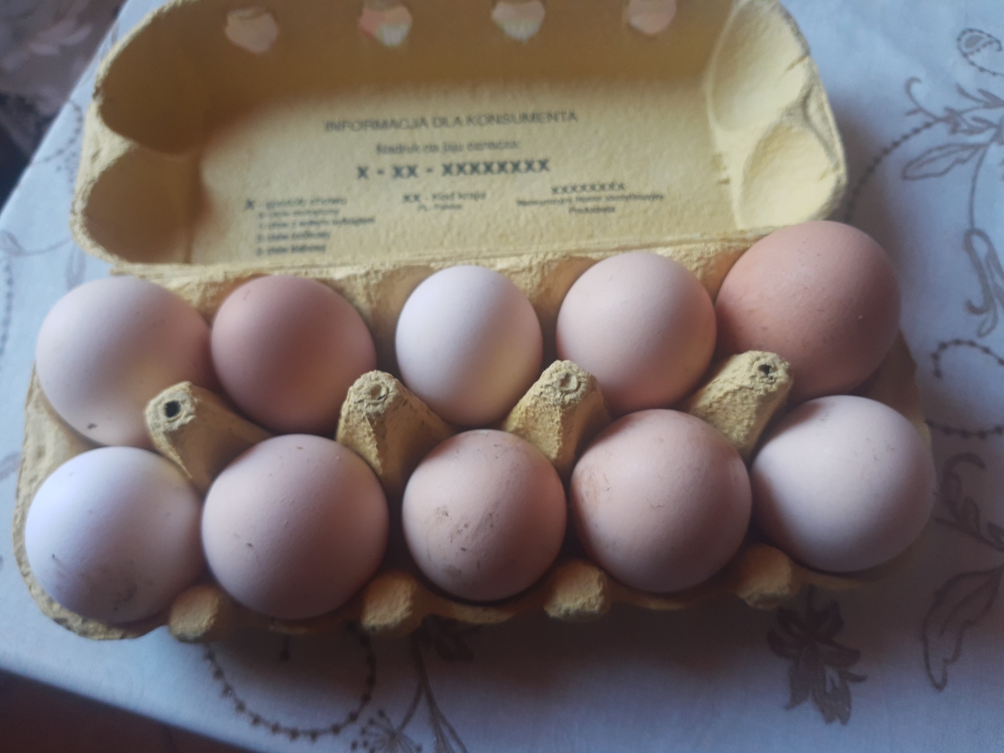 Otwarty karton z dziesięcioma jajami o różnych odcieniach brązu i bieli, widoczne zabrudzenia na niektórych skorupkach, karton z nadrukowanymi informacjami dla konsumenta, leżący na kwiecistej...