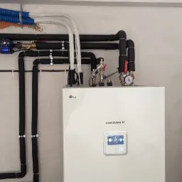 Biała pompa ciepła LG Therma V z czarnymi rurami instalacji grzewczej, widoczna skrzynka elektryczna i pakiet izolacji Pineltech przy ścianie.