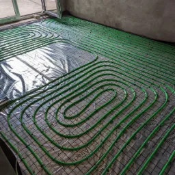 Zielone rurki ogrzewania podłogowego ułożone na czarnej siatce na folii, widok z góry, surowy beton na ścianie w tle.