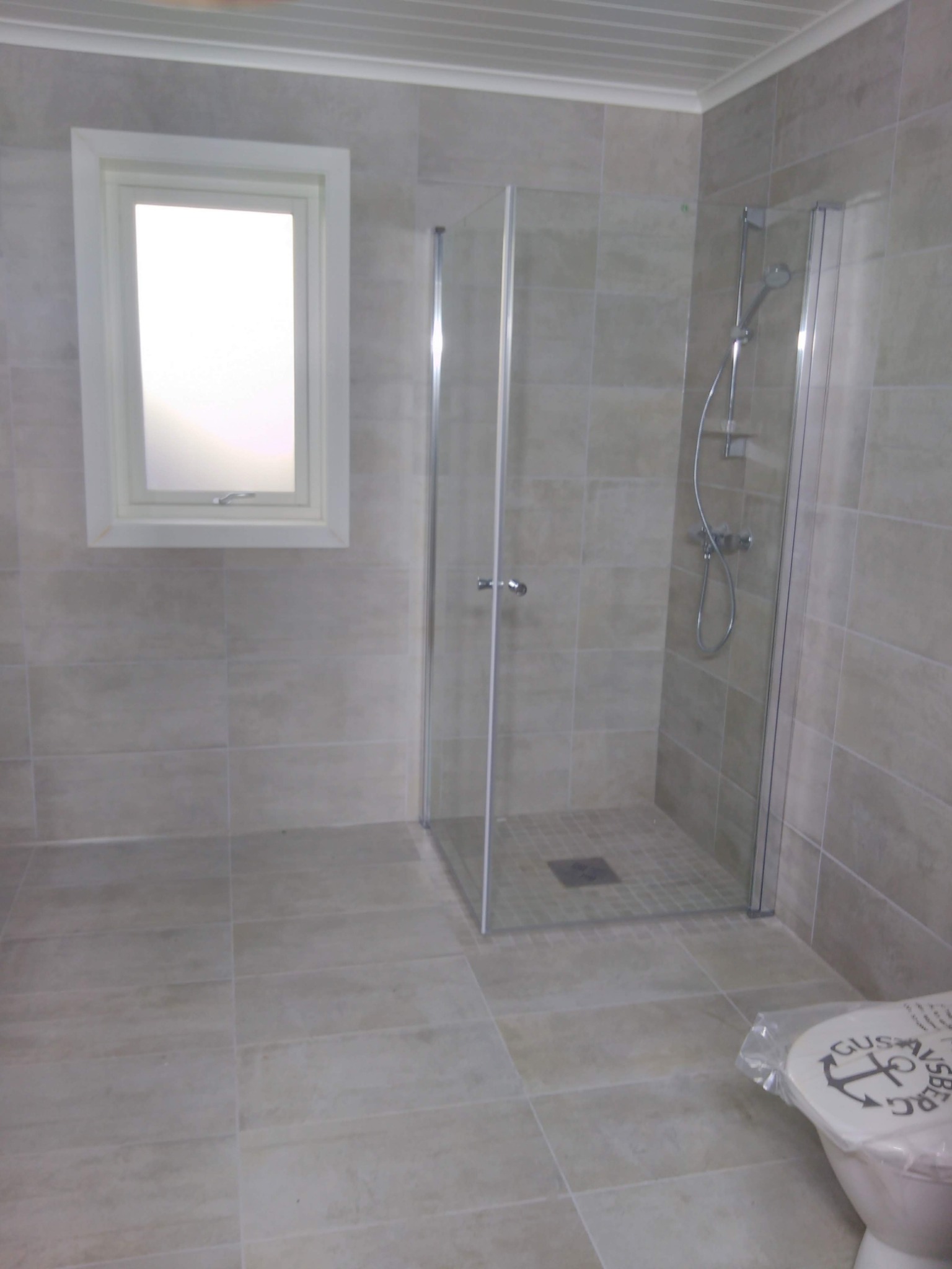 Wykończona łazienka w beżowych płytkach z kabiną prysznicową ze szklanymi drzwiami, widoczne okno z białymi ramami i toaleta z naklejoną folią ochronną na desce.