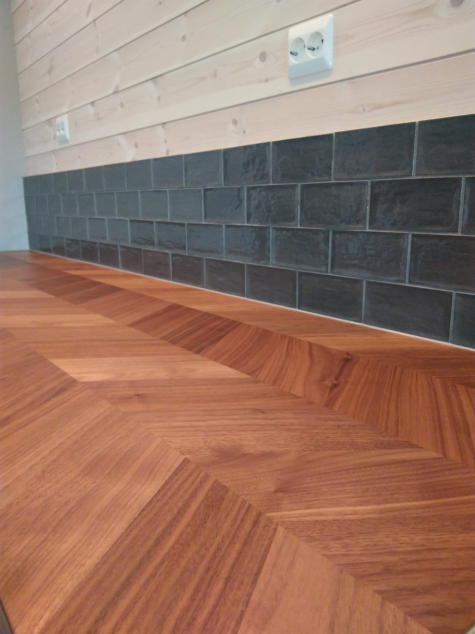 Ułożona w jodełkę drewniana powierzchnia blatu sąsiaduje z ciemnymi, prostokątnymi płytkami na ścianie i jasnymi drewnianymi panelami powyżej, z widocznym gniazdkiem elektrycznym.