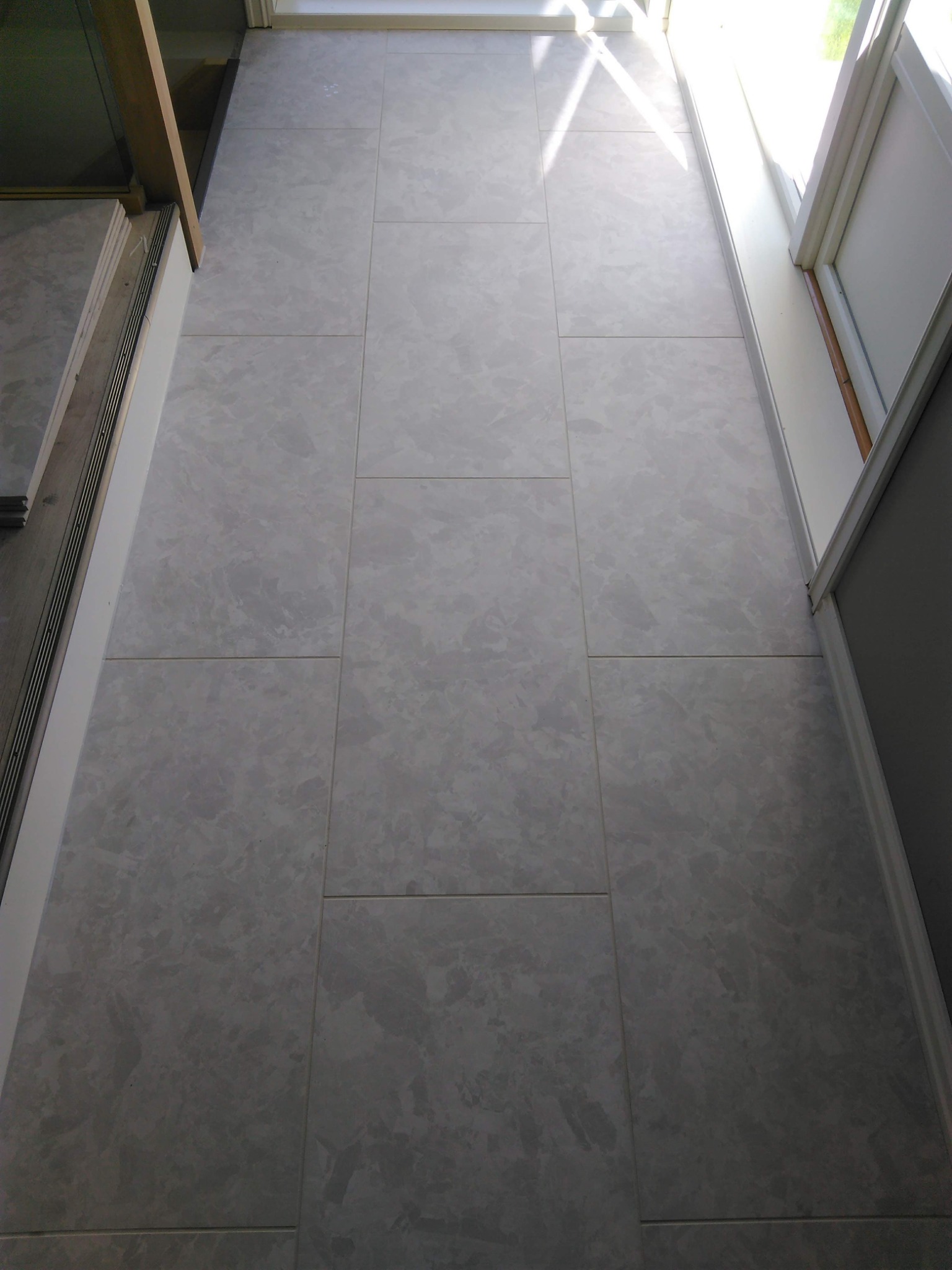 Ułożone jasnoszare płytki podłogowe z delikatnym wzorem marmurkowym, widoczne fugi, fragment progu i okna z promieniami słonecznymi.