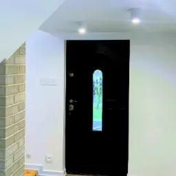 Wnętrze korytarza z czarnymi drzwiami wejściowymi z szybką, ceglanym obłożeniem filaru i trzema okrągłymi lampami sufitowymi.