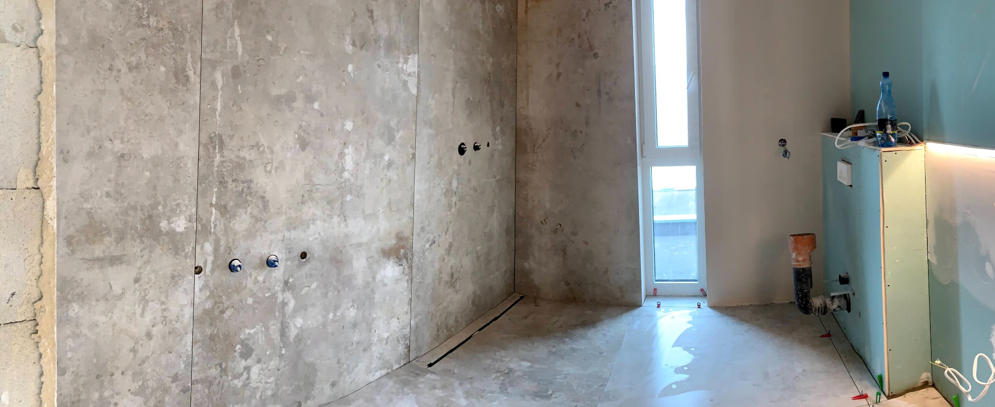 Surowa ściana z widocznymi instalacjami wodno-kanalizacyjnymi, fragment łazienki w trakcie remontu, z widoczną zabudową z płyt gipsowo-kartonowych i oknem w tle.