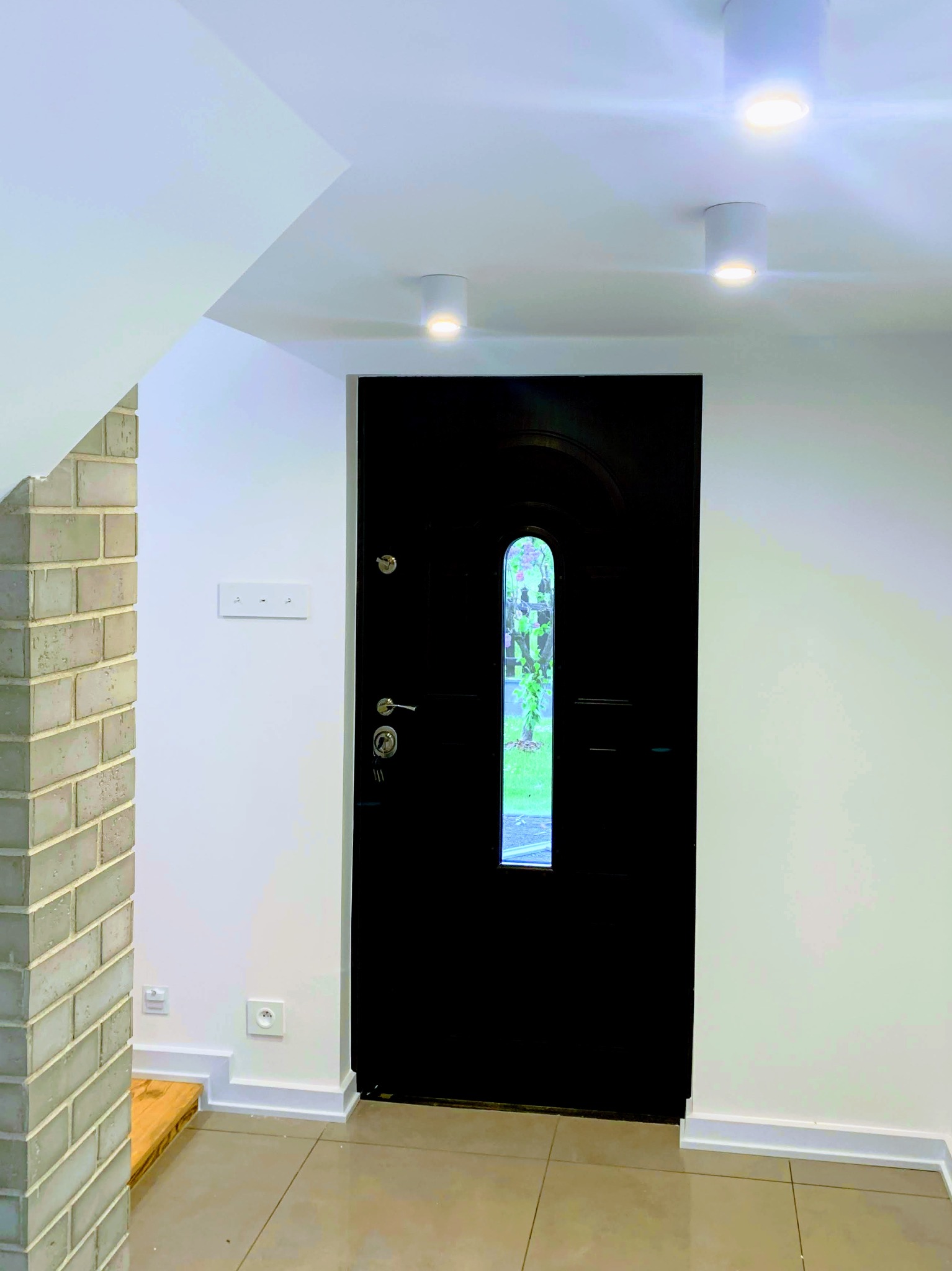Wnętrze korytarza z czarnymi drzwiami wejściowymi z szybką, ceglanym obłożeniem filaru i trzema okrągłymi lampami sufitowymi.
