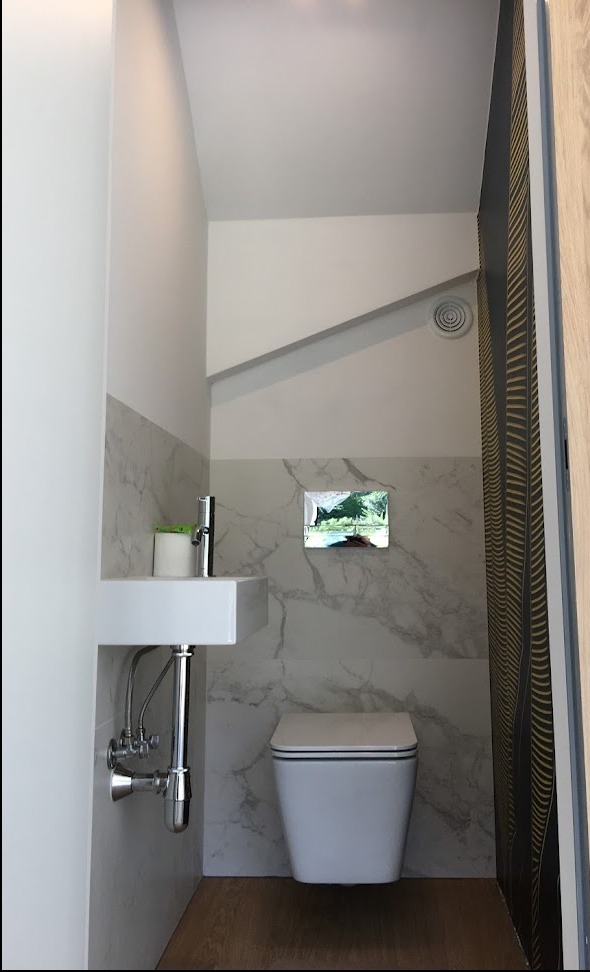 Nowoczesna, mała łazienka z marmurowymi płytkami, wiszącą toaletą z czarnym obramowaniem i kwadratową umywalką z chromowaną armaturą, uzupełniona drewnopodobną podłogą i wentylacją sufitową.