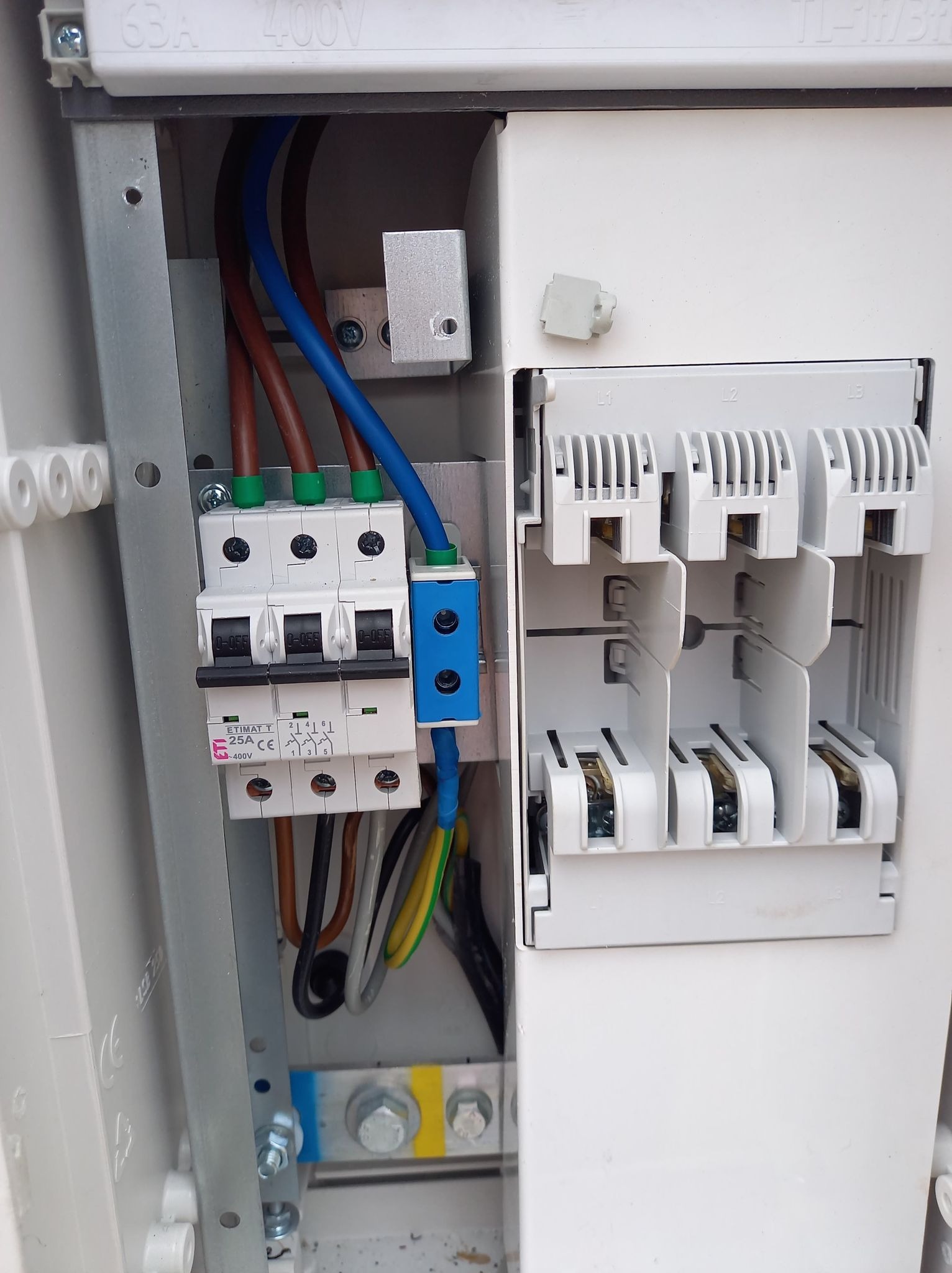 Otwarta skrzynka elektryczna z widocznymi bezpiecznikami, przewodami o różnych kolorach (brązowy, niebieski, żółto-zielony) i szyną wyrównawczą. Widoczne oznaczenia na bezpiecznikach i obudowie...