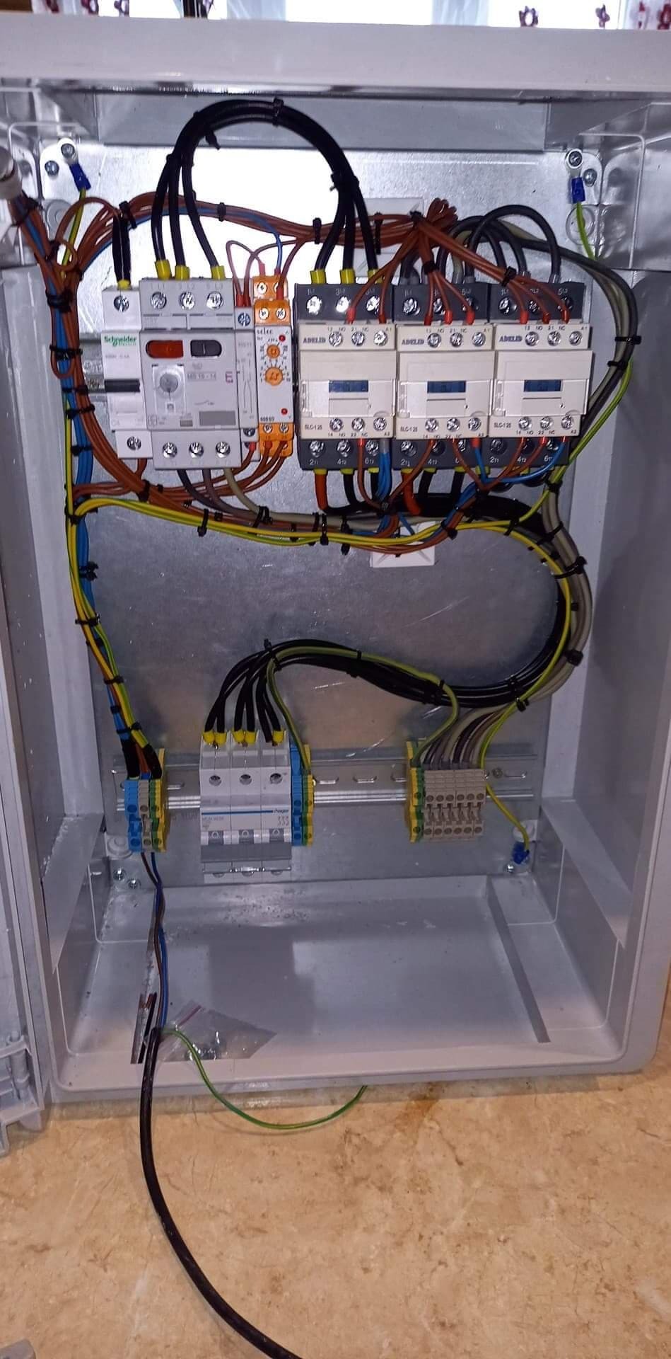 Wnętrze skrzynki elektrycznej z zamontowanymi stycznikami, bezpiecznikami i przewodami różnej grubości oraz kolorów, ułożonymi wewnątrz metalowej obudowy.