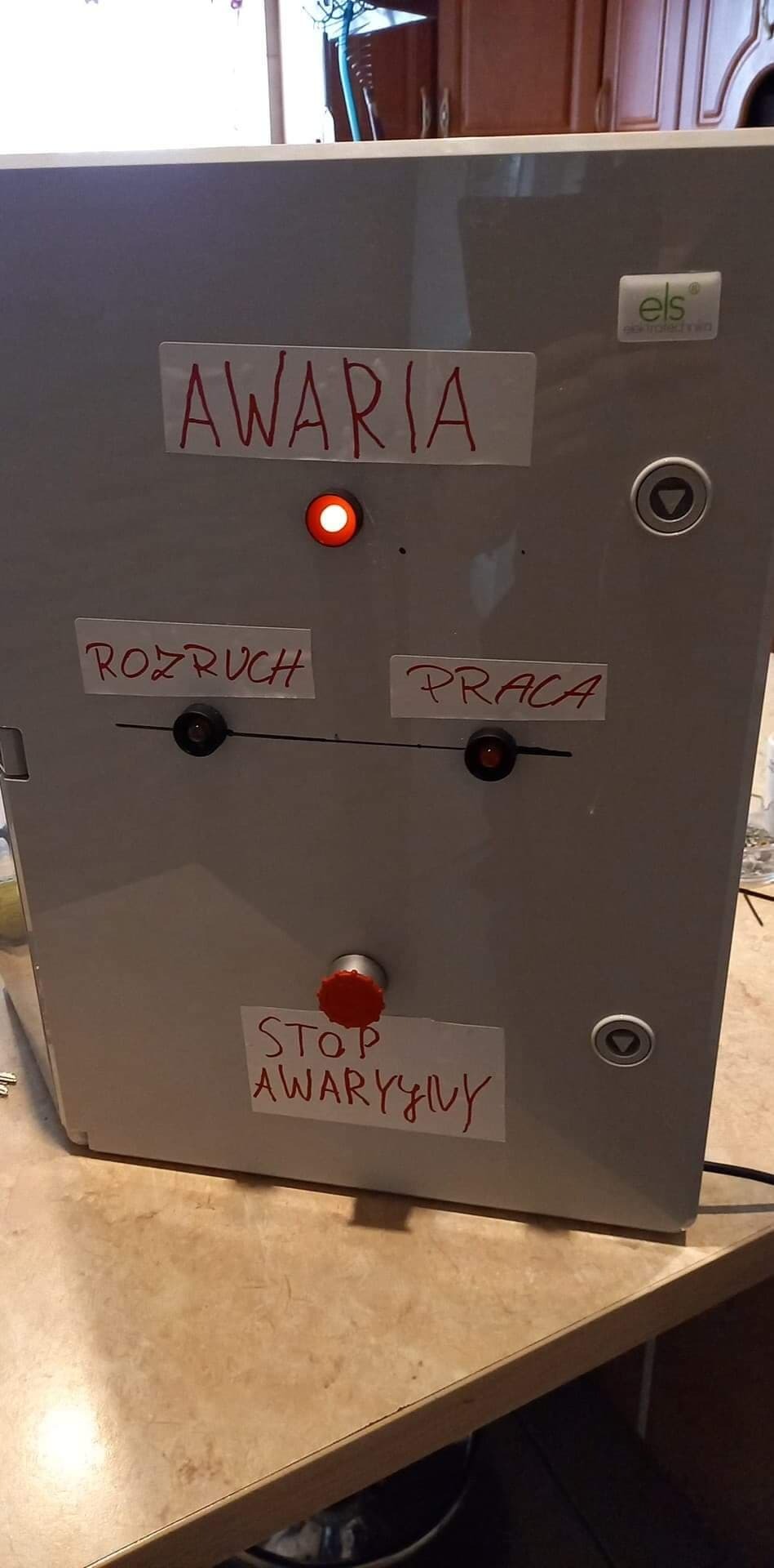Szara szafa sterownicza z napisem 'AWARIA' i czerwoną lampką sygnalizacyjną, przycisk 'STOP AWARYJNY' i etykiety 'ROZRUCH' i 'PRACA' z diodami, widoczne logo 'els elektrotechnika'.