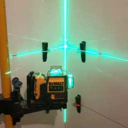 Laser krzyżowy DeWalt wyświetlający zielone linie na białych płytkach, z czarnymi klipsami poziomującymi, widoczne narożniki pomieszczenia.