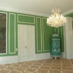 Jasnozielone wnętrze z dekoracyjnym piecem kaflowym, kryształowym żyrandolem i parkietem w Koszęcinie. Na ścianie obraz w ozdobnej ramie.