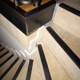 Detal wykończenia schodów z czarnymi antypoślizgowymi paskami, białą boazerią i metalową poręczą z drewnianym blatem.