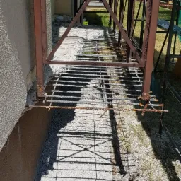 Metalowe rusztowanie przy budynku, widoczne ślady korozji na elementach konstrukcyjnych, cień rusztowania na ziemi.