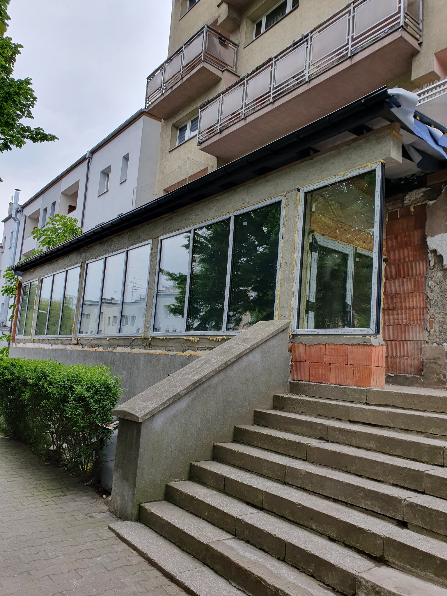Montaż dużych okien w surowym stanie budynku, widoczne cegły i betonowe schody wejściowe. Okna z czarnymi ramami odbijają otoczenie.