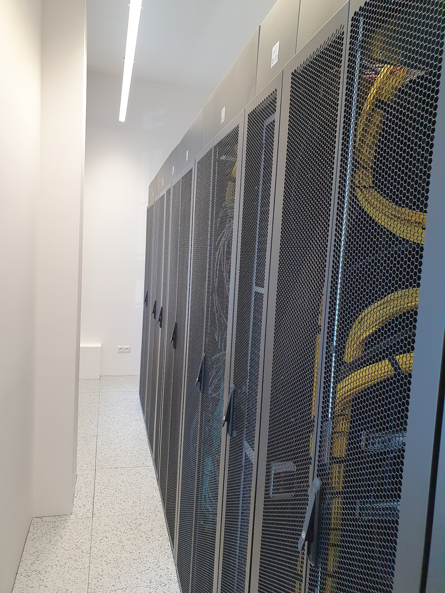 Rząd szaf serwerowych z perforowanymi drzwiami w chłodnym, białym pomieszczeniu z terakotą, widoczne kable wewnątrz szaf.